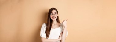 Gözlüklü, gülümseyen ve solu işaret eden mutlu kadın, boş alanda logo gösteriyor, bej arka planda duruyor..