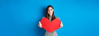 Sevgililer günü ve ilişki konsepti. Güzel romantik kız seni sevdiğimi söylüyor, büyük kırmızı bir kalple elini uzatarak, mavi arka planda durarak..