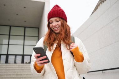 Happy girl student in red hat, holds smartphone, tourist looks at map app on her phone, explores sightseeing, texts message, looks for couchsurfing, rents place to stay online.