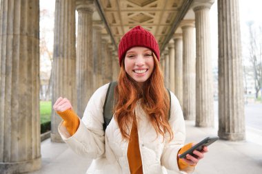 Mobile broadband and people. Smiling redhead 20s girl with backpack, uses smartphone on street, holds mobile phone and looks at application.
