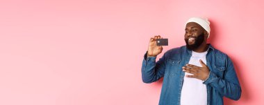 Alışveriş konsepti. Mutlu Afro-Amerikalı bir adamın resmi kredi kartından memnun görünüyor, bankayı tavsiye ediyor, pembe arka planda duruyor..