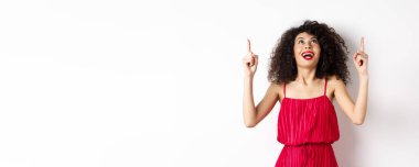 Heyecanlı beyaz kadın kıvırcık saçlı, kırmızı dudaklı, şaşkın bir gülümsemeyle bakıyor ve işaret ediyor, tanıtım teklifini kontrol ediyor, beyaz arka planı var..