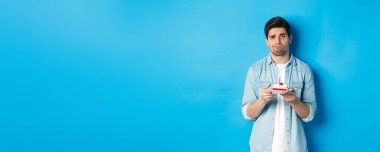 Üzgün ve kasvetli bir adam doğum günü pastası tutuyor, somurtuyor, mavi arka planda duruyor..