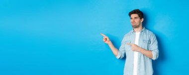 Şüpheli yetişkin bir adam terfiyi sol eliyle işaret ediyor ve kararsız görünüyor, hayal kırıklığına uğruyor, mavi arka planda duruyor..