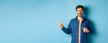 Tişörtlü yakışıklı genç adam, solu işaret ediyor, logoyu gösteriyor, mavi arka planda duruyor..