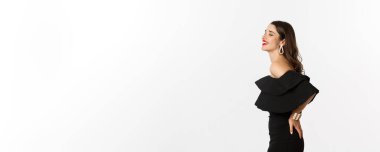 Siyah parti elbisesi içinde duran, gülen, kaygısız, beyaz arka planda gülümseyen çekici bir kadın modelin profil görüntüsü. Boşluğu kopyala