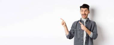 Yakışıklı, gülümseyen adam reklama bakıyor, bakıyor ve solu işaret ediyor, heyecanlı yüzüyle, beyaz arka planda duruyor..