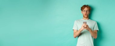 Kızıl saçlı, gözlüklü, elinde cep telefonu olan etkileyici bir genç adam kameraya hayret ediyor ve bakıyor..