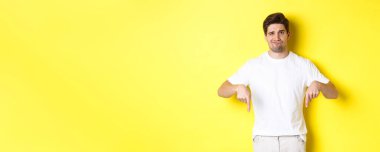 Genç mutsuz adam yüzünü buruşturuyor, reklamları işaret ediyor, üründen hayal kırıklığına uğruyor, sarı arka planda duruyor..