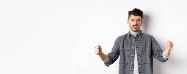 Şüpheci genç adam büyük bir nesne gösteriyor ve somurtuyor, beyaz arka planda isteksiz duruyor..