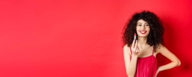 Kıvırcık saçlı, kırmızı ruj sürmüş, gülümseyen, kozmetik ürünler öneren, beyaz arka planda duran....