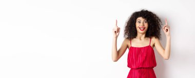 Kırmızı elbiseli ve makyajlı mutlu zarif bir kadın, gülümsüyor ve logoyu işaret ediyor, reklamını yapıyor, beyaz arka planda duruyor..