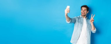 Mutlu adam selfie çekiyor ve mavi arka planda barış işareti gösteriyor, elinde cep telefonu var..