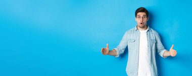 Şaşkın yakışıklı adam elinde büyük bir şey tutuyor, büyük boy ve şaşkın bir yüz gösteriyor, mavi arka planda duruyor..