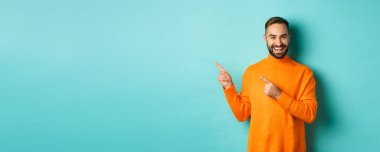 Yakışıklı, gülümseyen adam parmaklarını sola gösteriyor, logonuzu gösteriyor, turkuaz süveter giymiş, turkuaz arka planda duruyor..
