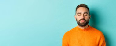 Süveter giyen, dudaklarını büzen, gözlerini kapayan ve öpücüğü bekleyen, açık mavi arka planda duran genç adamın yakın çekimi..