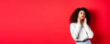 Kırmızı dudaklı güzel ve şık kıvırcık kadın, mükemmel genç yüze dokunuyor ve kameraya şehvetli bakıyor, kırmızı arka planda duruyor..