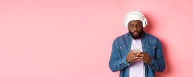 Adam telefonda mesajları okurken ciddi görünüyor, kulaklıkla müzik dinliyor, pembe arka planda duruyor..