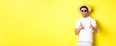 Tatilde seninle flört eden, kamerayı işaret eden ve göz kırpan güneş gözlüklü, sarı arka planda yaz şapkası takan kendinden emin ve küstah bir adam..