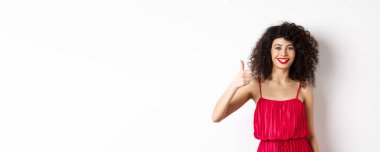 Kırmızı dudaklı ve zarif elbiseli neşeli bir kadın. Baş parmağını kaldırıyor ve gülümsüyor. Ürün öneriyor, beyaz arka planda duruyor..