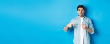 Çevrimiçi alışveriş, uygulama ve teknoloji kavramı. Etkileyici bir adam cep telefonunu işaret ediyor ve çok şaşırmış görünüyor. Uygulama öneriyor, mavi arka planda duruyor..