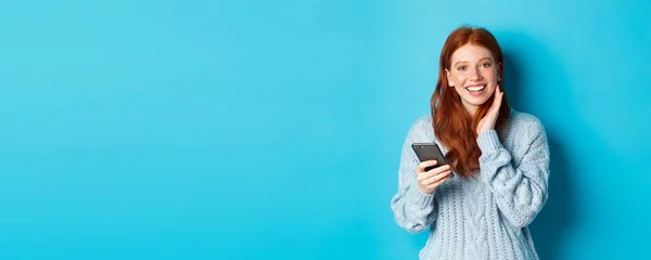 Beautiful redhead girl in sweater, smiling at camera, using mobile phone app, standing with smartphone against blue background.
