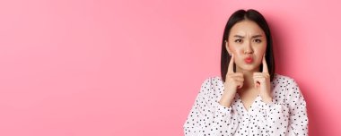 Güzellik ve yaşam tarzı konsepti. Şirin Asyalı kadın somurtkan surat yapıyor, yanakları dürtüyor ve yüzünü buruşturuyor, pembe arka planda duruyor..
