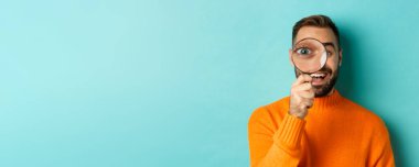 Komik adam büyüteçten bakıyor, bir şeyi arıyor ya da araştırıyor turkuaz arka planda turuncu süveter giyiyor..