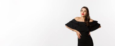 Moda ve güzellik. Siyah elbiseli, makyajlı, kırmızı dudaklı, beyaz arka planda kendine güvenen, beyaz arka planda duran modern bir kadın..