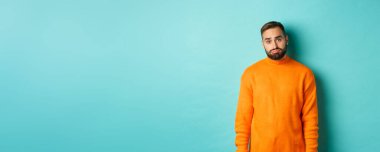 Üzgün adam kameraya bakıyor, somurtuyor ve sıkıntılı hissediyor, açık mavi arka planda duruyor..