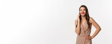 Noel partisi ve kutlama konsepti. Kırmızı dudaklı çekici genç bir kadın, lüks elbiseler giyiyor, cep telefonuyla konuşuyor ve gülümsüyor, beyaz arka planda duruyor..