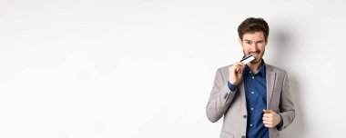 Başarılı takım elbiseli genç işadamı kredi kartını ısırıyor ve beyaz arka planda arsız görünüyor..