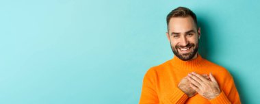 Yakışıklı delikanlının yakın çekimi, teşekkür ediyor, kalbi kucaklıyor ve gülümsüyor, açık mavi arka planda duruyor..