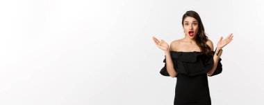 Güzellik ve moda konsepti. Heyecanlı güzel bir kadın şaşkınlıkla bakıyor, iyi habere tepki veriyor, siyah elbise içinde makyajlı, beyaz arka planda..