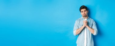 Yardım için yalvaran, somurtan ve üzgün görünen genç bir adam bir şey istiyor, mavi arka plana karşı duruyor..