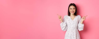 Güzel Asyalı kadın alışverişte seçim yapıyor, parmaklarını yana doğru gösteriyor ve değişkenleri gösteriyor, kameraya gülümsüyor, pembe arka planda duruyor..