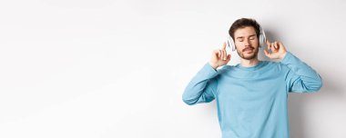 Rahatlamış genç adam favori şarkının tadını çıkarıyor, kulaklıkla müzik dinliyor, gözleri kapalı ve sakin bir yüz, beyaz arka planda duruyor..