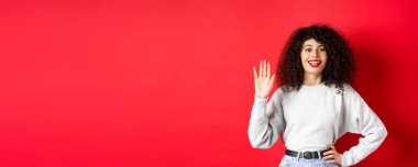Kıvırcık saçlı arkadaş canlısı kadın seni selamlıyor, elini sallıyor ve merhaba diyor, kendini tanıtıyor, kırmızı arka planda duruyor..