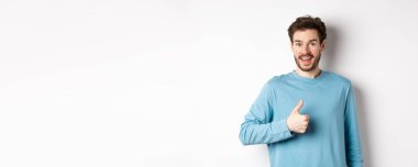 Neşeli genç adam kabul ediyor ve kabul ediyor ve evet diyor, onaylanmış olarak baş parmağını göster, kameraya mutlu bir şekilde gülümse, beyaz arka planda dur.