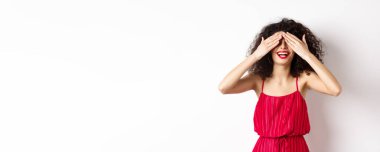 Heyecanlı kıvırcık saçlı, parti elbisesi giymiş, gözleri kapalı, kırmızı dudaklarıyla gülümseyen, mutlu yüzüyle şaşırmayı bekleyen, beyaz arka planda duran.