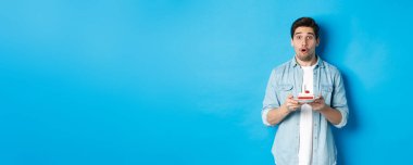 Doğum günü pastası tutan ve şaşırmış görünen bir adam, yanan mumun üzerinde bir dilek diliyor, mavi arka planda duruyor..