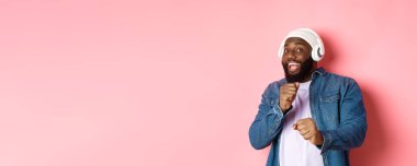 Mutlu Afro-Amerikalı adam dans ediyor, kulaklıkla müzik dinliyor ve pembe arka planda kameraya bakıyor..