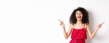 Kırmızı elbiseli mutlu güzel bayan, gülüyor ve yandan işaret ediyor, iki yol gösteriyor, beyaz arka planda duruyor..