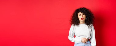 Kadın hasta, eğiliyor ve karnına dokunuyor, karın ağrısı ya da regl sancısı çekiyor, kırmızı arka planda duruyor..