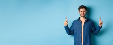 Şüpheci genç adam kaşlarını çatmış, gözlerini kısmış, iğrenç bir şeyden korkup mavi arka planda duruyor..