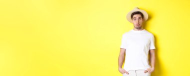 Hasır şapkalı kafası karışmış gezgin kafası karışmış, kameraya bakıyor, sarı arka planda dikiliyor. Boşluğu kopyala