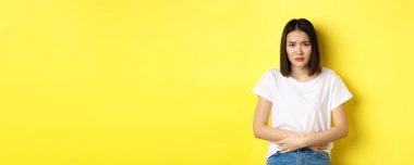 Kendini hasta hisseden Asyalı bir kadın tasviri, ağrılı kramplar, karın ağrısı ve acıdan kaşlarını çatma, regl döneminde rahatsızlık, sarı arka planda dikilme..