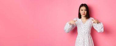 Hayal kırıklığına uğramış Asyalı kadın manken tanıtım teklifini işaret ediyor. Kaşlarını çatıyor ve kameraya bakıyor. Pembe arka planda dikiliyor..