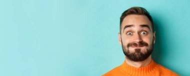 Komik sakallı adamın suratını gösteren, somurtan, nefes alan turuncu süveter giymiş turkuaz arka planda dikilen fotoğrafı..