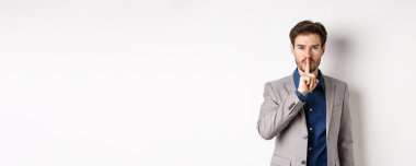 Yakışıklı maço takım elbiseli adam kameraya şşş yapıyor, parmak dudağa basıyor, sesini alçalt, sırrını söyle, beyaz arka plana karşı duruyor..